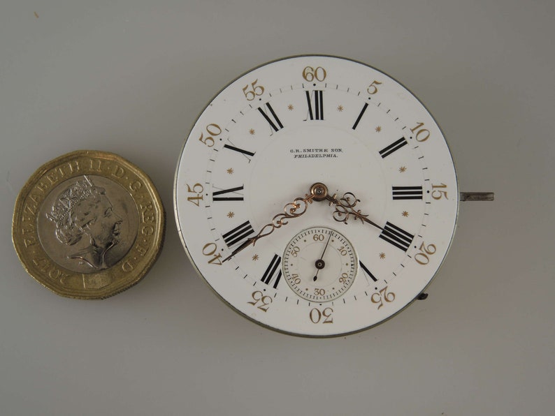 May include: A white pocket watch face with black Roman numeral hour markers and gold Arabic numeral minute markers. The watch face has a smaller, circular sub-dial with black markings and a gold hand. The watch face is signed "G.R. Smith & Son, Philadelphia."