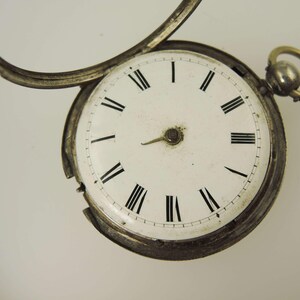 May include: Close-up of an antique pocket watch with a white face and black Roman numerals. The watch has a silver-colored metal casing and a small loop at the top. The watch's hands point to approximately 1:50.