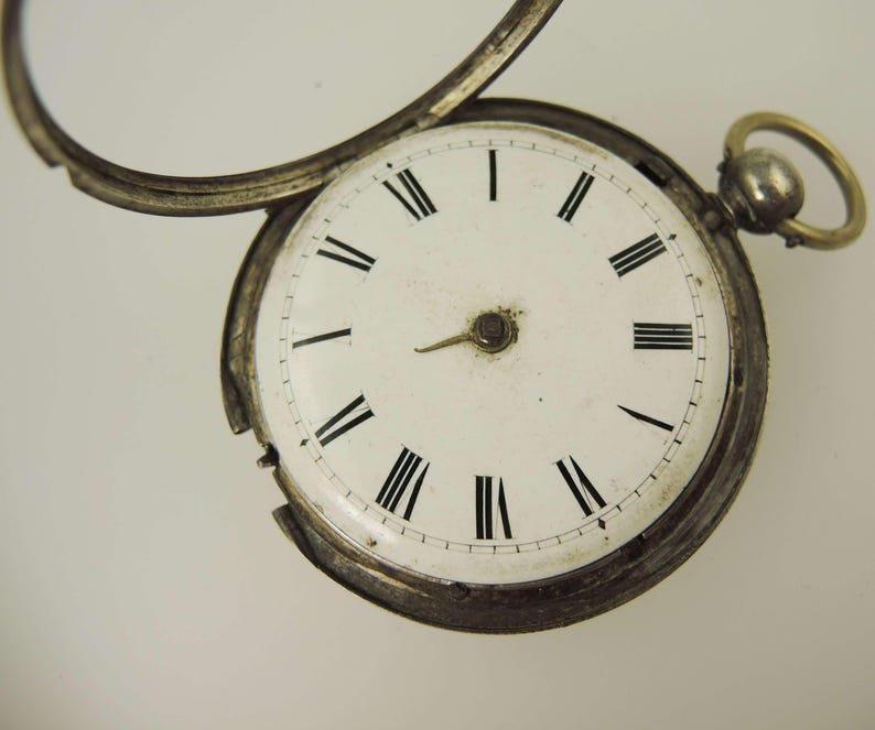 May include: Close-up of an antique pocket watch with a white face and black Roman numerals. The watch has a silver-colored metal casing and a small loop at the top. The watch's hands point to approximately 1:50.