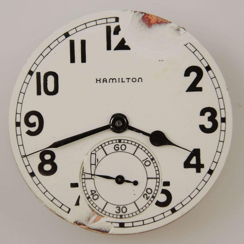May include: A vintage Hamilton watch face with a white background, black numerals, and black hands. The watch face has a subdial and the brand name "HAMILTON" is printed at the top. The face shows signs of wear and tear.