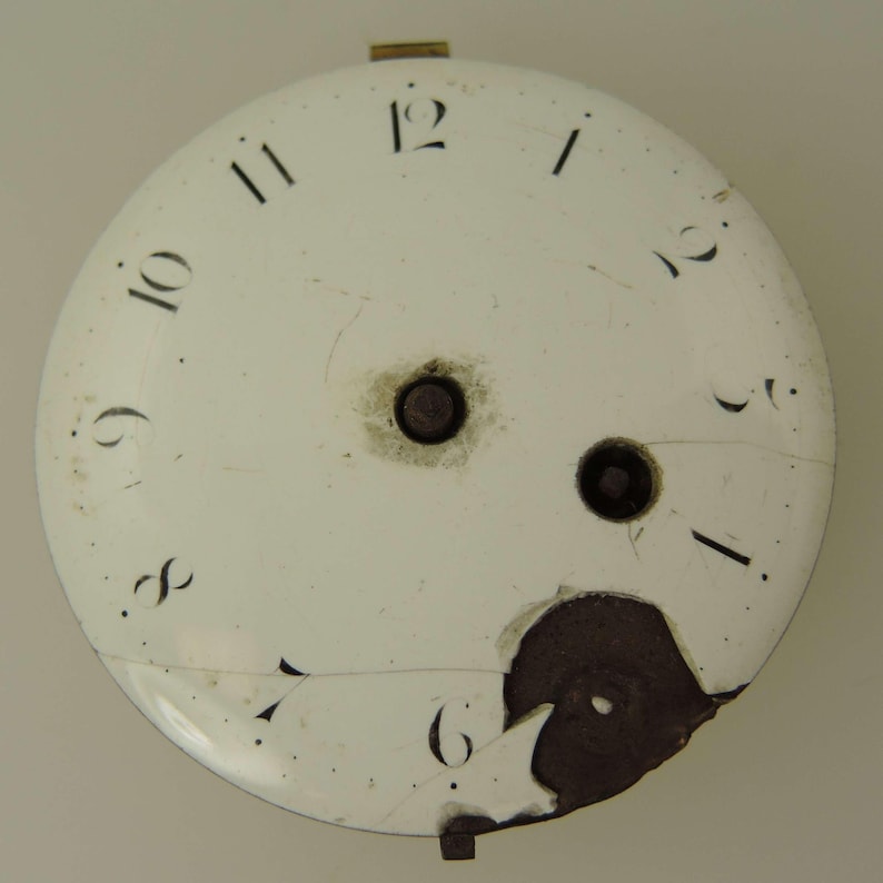 Puede incluir: Una esfera de reloj blanca antigua con n&uacute;meros negros. La esfera est&aacute; agrietada y da&ntilde;ada, con dos agujeros centrales y una zona marr&oacute;n oscura donde el esmalte se ha desprendido. La esfera del reloj tiene un peque&ntilde;o accesorio met&aacute;lico en la parte superior e inferior.