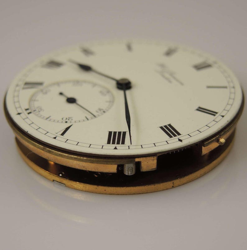 May include: A close-up of a white pocket watch face with black Roman numeral hour markers and black hands. The watch has a gold-toned case and a small seconds dial at the 12 o'clock position.