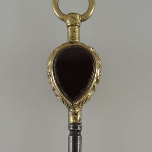 May include: A gold-toned watch key with a teardrop-shaped red stone set in a decorative frame. The key has a loop at the top for attaching to a chain.