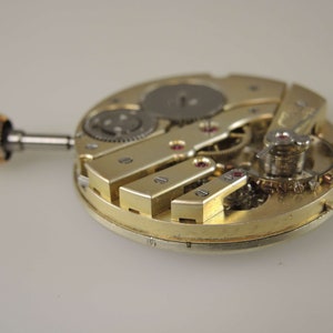 Puede incluir: Primer plano de un movimiento de reloj de tono dorado con engranajes intrincados y un v&aacute;stago de cuerda. El movimiento est&aacute; expuesto, mostrando el funcionamiento interno del reloj.