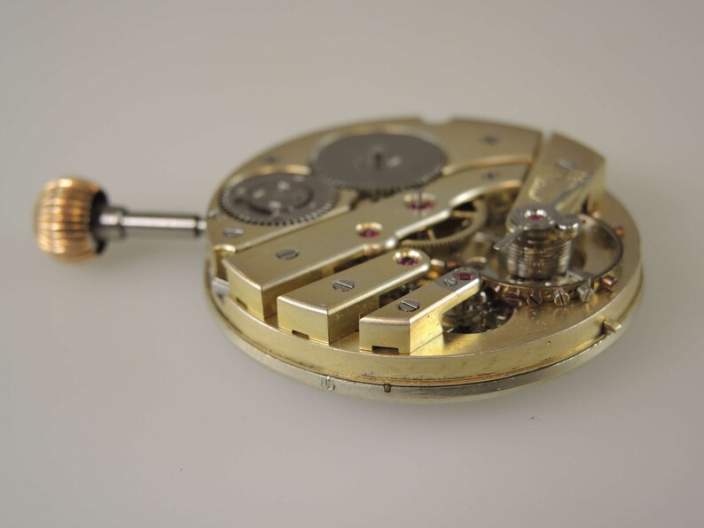 Puede incluir: Primer plano de un movimiento de reloj de tono dorado con engranajes intrincados y un v&aacute;stago de cuerda. El movimiento est&aacute; expuesto, mostrando el funcionamiento interno del reloj.