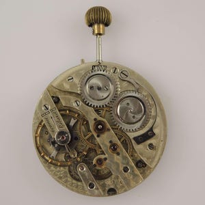 May include: A detailed view of a vintage watch movement, showcasing intricate gears, springs, and a balance wheel. The mechanism is primarily silver-toned with brass accents, and a winding stem with a textured, gold-colored crown.
