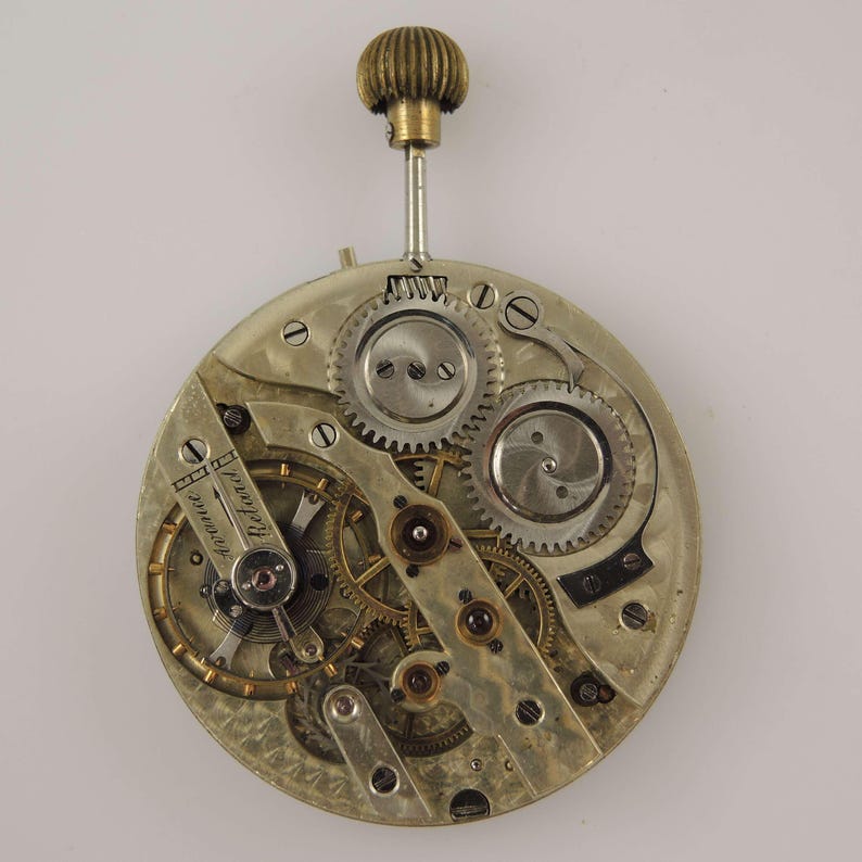 May include: A detailed view of a vintage watch movement, showcasing intricate gears, springs, and a balance wheel. The mechanism is primarily silver-toned with brass accents, and a winding stem with a textured, gold-colored crown.