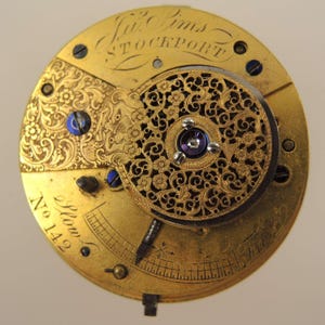 May include: Close-up of a vintage watch movement with intricate gold filigree and floral designs. The watch mechanism features engraved text, including the name "Jno. Pims STOCKPORT" and the number "142". The movement has blue screws and a dark center.
