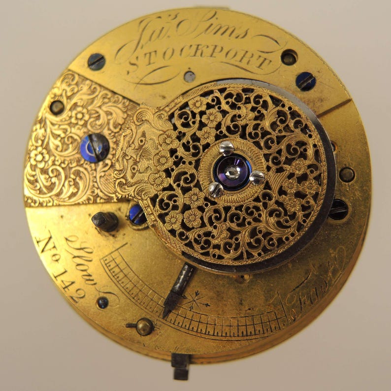 May include: Close-up of a vintage watch movement with intricate gold filigree and floral designs. The watch mechanism features engraved text, including the name "Jno. Pims STOCKPORT" and the number "142". The movement has blue screws and a dark center.