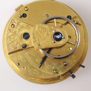 May include: Close-up of a vintage gold pocket watch movement. The intricate mechanism features engraved floral patterns, a balance wheel, and a dark blue jewel. The watch's hands are visible, and the overall design is ornate and detailed.