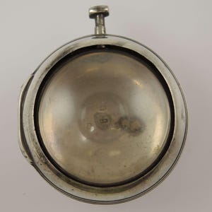 Könnte beinhalten: Eine silberne Taschenuhr mit kreisförmigem Zifferblatt und einem Stiel oben. Die Uhr hat eine polierte Oberfläche und eine detaillierte Lünette. Das Zifferblatt ist sichtbar, und das Innere hat ein leicht angelaufenes Aussehen. Eine klassische Uhr.
