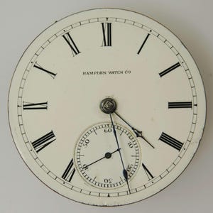 Puede incluir: Esfera de reloj de bolsillo antiguo con fondo color crema. Los n&uacute;meros romanos negros marcan las horas, con un dial de segundos m&aacute;s peque&ntilde;o en la parte inferior. Las palabras "HAMPDEN WATCH CO" est&aacute;n impresas en la parte superior. Las manecillas son negras y la esfera tiene un borde met&aacute;lico.
