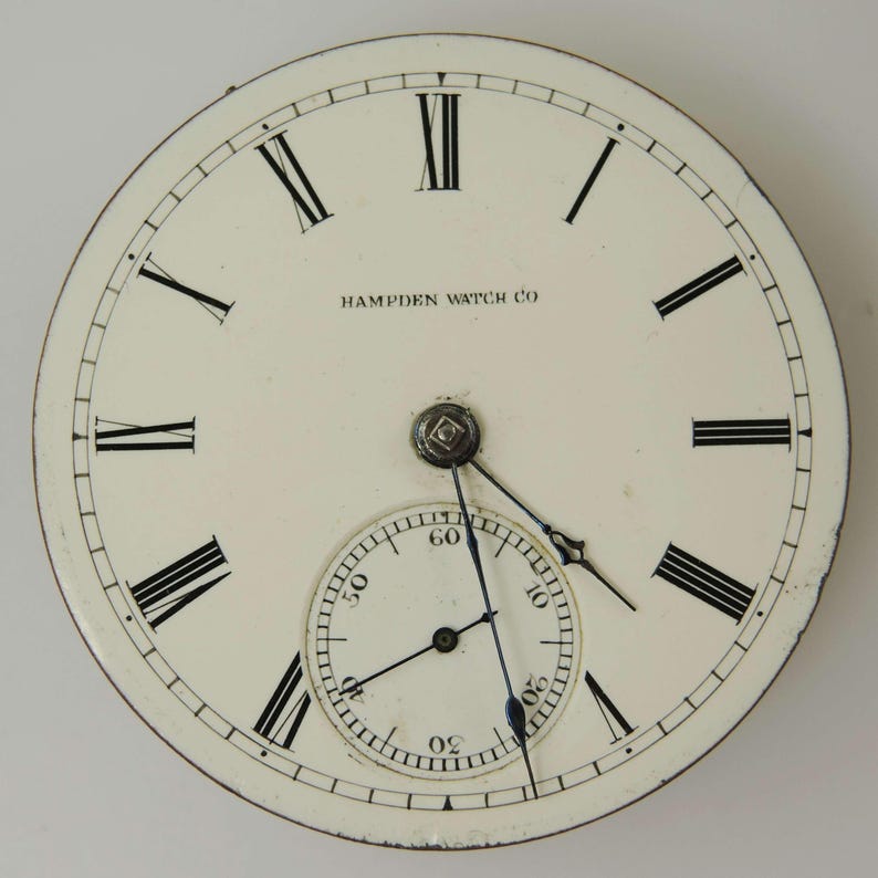 Puede incluir: Esfera de reloj de bolsillo antiguo con fondo color crema. Los n&uacute;meros romanos negros marcan las horas, con un dial de segundos m&aacute;s peque&ntilde;o en la parte inferior. Las palabras "HAMPDEN WATCH CO" est&aacute;n impresas en la parte superior. Las manecillas son negras y la esfera tiene un borde met&aacute;lico.