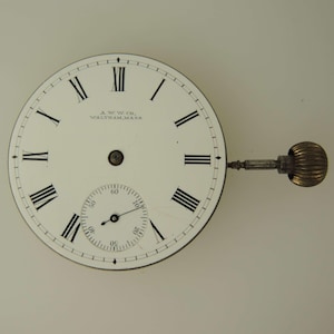 May include: A vintage pocket watch face with a white background, featuring black Roman numerals and a smaller seconds dial. The watch face has a stem and a textured, gold-colored winding crown. The text "A.W.W. Co. Waltham, Mass." is printed on the face.