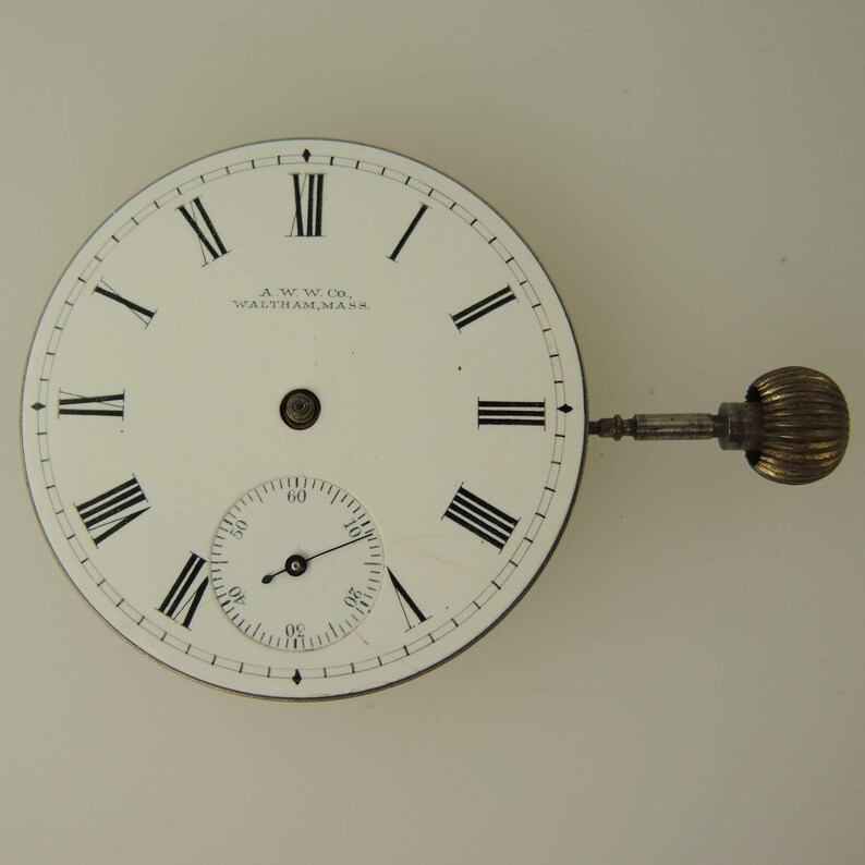 May include: A vintage pocket watch face with a white background, featuring black Roman numerals and a smaller seconds dial. The watch face has a stem and a textured, gold-colored winding crown. The text "A.W.W. Co. Waltham, Mass." is printed on the face.