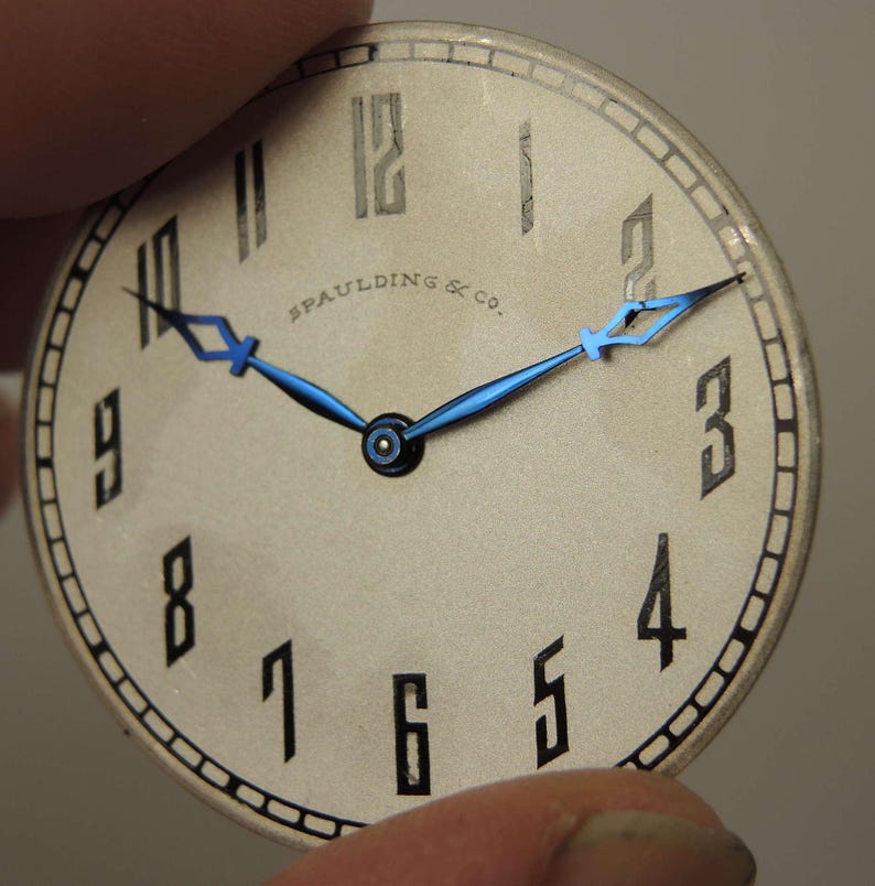May include: A white watch face with black Roman numeral hour markers and black Arabic numeral minute markers. The watch face has a blue steel hour and minute hand. The text "SPAULDING & CO." is printed on the watch face.