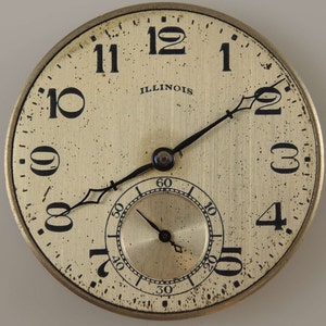 Op de afbeelding: Een vintage Illinois zakhorloge met zwarte wijzers en Romeinse cijfers voor de uren. De wijzerplaat van het horloge is cr&egrave;mekleurig met een goudkleurige subdial.