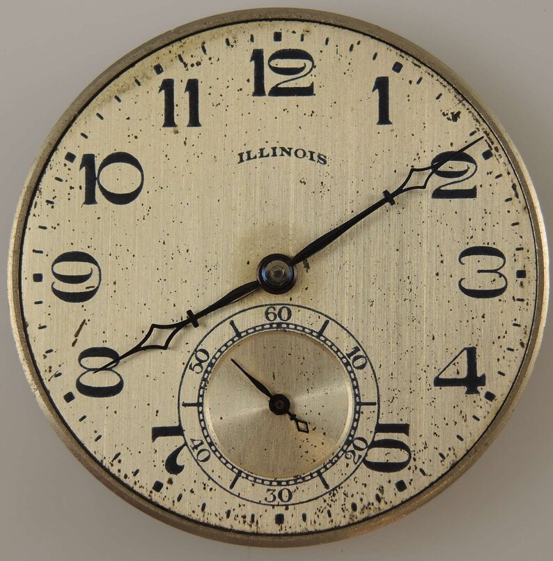 Op de afbeelding: Een vintage Illinois zakhorloge met zwarte wijzers en Romeinse cijfers voor de uren. De wijzerplaat van het horloge is cr&egrave;mekleurig met een goudkleurige subdial.