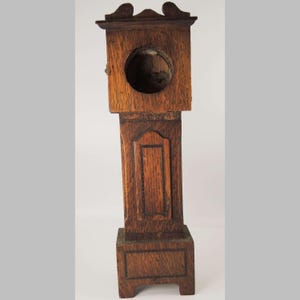 May include: A miniature wooden grandfather clock with a dark brown finish. The clock has a rectangular base, a long, detailed body, and a top with a decorative curved design. The clock face is a circular opening. The clock is a vintage item.