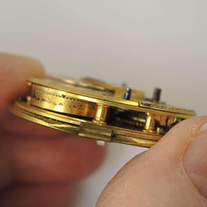 Pode incluir: Close-up de um movimento de rel&oacute;gio vintage, mostrando engrenagens e componentes dourados intrincados. O mecanismo do rel&oacute;gio &eacute; segurado em uma m&atilde;o, revelando o artesanato detalhado do rel&oacute;gio antigo. A imagem destaca a precis&atilde;o e complexidade do funcionamento interno do rel&oacute;gio.