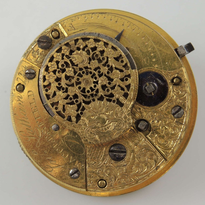 Op de afbeelding: Een gouden zakhorloge met ingewikkelde bloemmotieven en een gedetailleerd gezicht met Romeinse cijfers. Het horloge heeft een klein, rond, open gebied in het midden met een decoratief ontwerp.