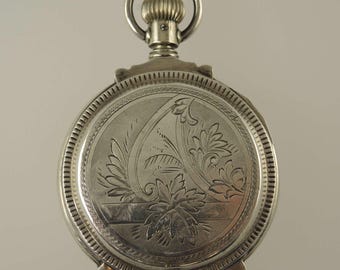 May include: A silver pocket watch with an engraved floral design on the cover. The watch has a round face with a detailed border and a loop at the top for a chain. The watch is a vintage item.