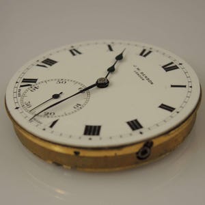 Puede incluir: Una esfera de reloj de bolsillo blanca con marcadores de hora en n&uacute;meros romanos negros y agujas negras. La esfera del reloj tiene el texto "J.W. Benson London" impreso en ella. El reloj est&aacute; en una caja de color dorado.