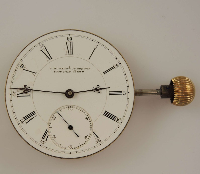 Puede incluir: Una esfera de reloj de bolsillo blanca con marcadores de hora en n&uacute;meros romanos negros y agujas negras. La esfera del reloj tiene un c&iacute;rculo conc&eacute;ntrico m&aacute;s peque&ntilde;o con marcas negras para los segundos. La esfera del reloj est&aacute; inscrita con "E. Howard & Co Boston Pat'd Feb 4th 1868".