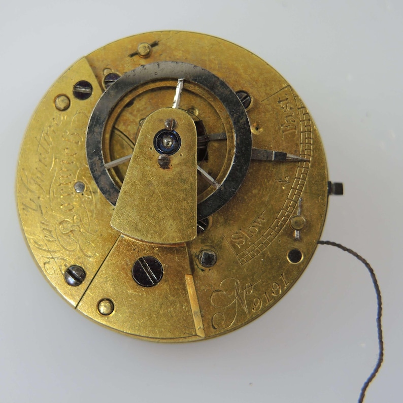 May include: Close-up of a vintage watch movement with gold-colored gears and a silver balance wheel. The watch mechanism has intricate details, including screws, a balance cock, and the inscription "Fast" and "Slow". The number "9191" is also visible.