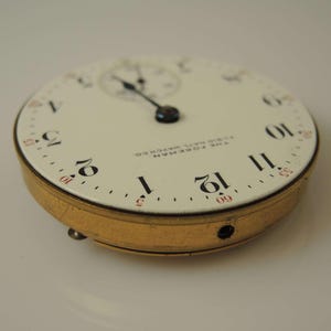 K&ouml;nnte beinhalten: Eine wei&szlig;e Taschenuhr mit schwarzen r&ouml;mischen Ziffern f&uuml;r die Stunden und schwarzen Zeigern. Das Zifferblatt der Uhr hat auch ein kleineres Zifferblatt mit einem schwarzen Zeiger. Das Zifferblatt der Uhr zeigt "The Foreman Fine Natl Watch Co."