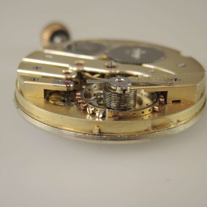 Puede incluir: Primer plano de un movimiento de reloj de tono dorado con engranajes y resortes intrincados. El reloj est&aacute; desmontado y se pueden ver las partes internas.