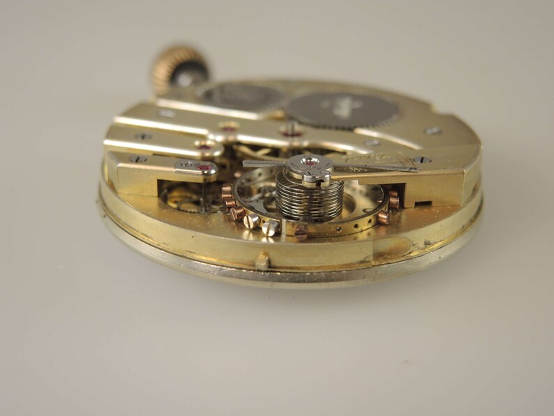 Puede incluir: Primer plano de un movimiento de reloj de tono dorado con engranajes y resortes intrincados. El reloj est&aacute; desmontado y se pueden ver las partes internas.