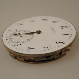 May include: A white pocket watch with black Roman numeral hour markers and a smaller sub-dial. The watch has a gold-tone case and is in a slightly open position.