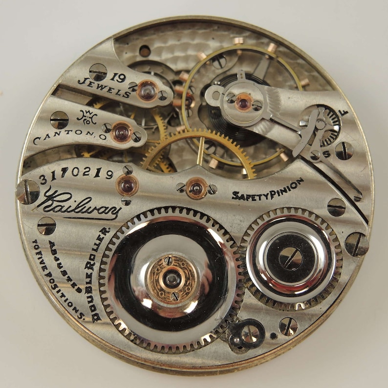K&ouml;nnte beinhalten: Eine Nahaufnahme des Innenlebens einer silbernen Taschenuhr. Das Zifferblatt der Uhr ist mit "Railway" und "Double Roller Adjusted to Five Positions" beschriftet. Die Uhr hat 19 Steine und ein Sicherheitszahnrad.