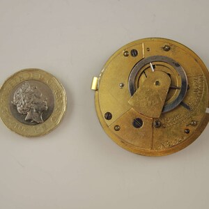 May include: Close-up of a gold-colored pocket watch movement with intricate gears and a silver-colored dial. The watch movement is visible through a clear glass cover. The watch is in a disassembled state, showing the inner workings of the mechanism.