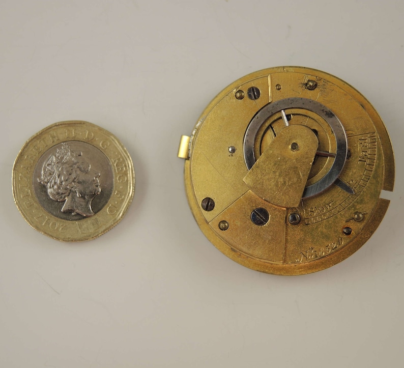 May include: Close-up of a gold-colored pocket watch movement with intricate gears and a silver-colored dial. The watch movement is visible through a clear glass cover. The watch is in a disassembled state, showing the inner workings of the mechanism.