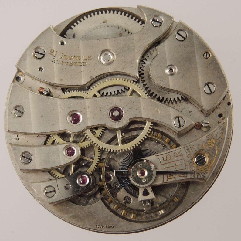 May include: Close-up of a vintage watch movement with visible gears, screws, and jewels. The text "21 Jewels Adjusted" is visible. The watch mechanism is silver-toned with intricate details, showcasing the internal workings of a timepiece.