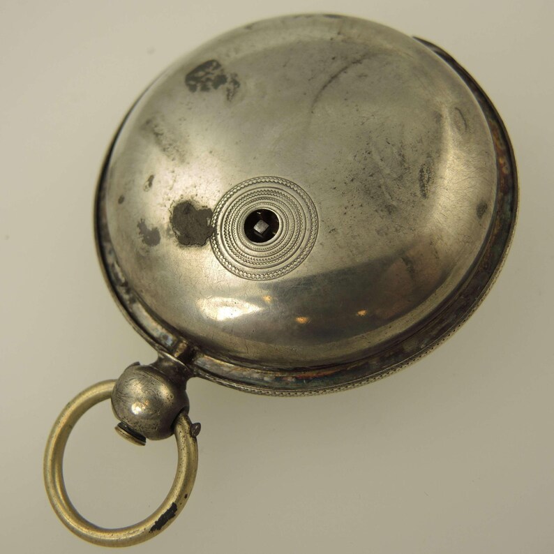 May include: A tarnished silver pocket watch with a circular design. The watch features a decorative ring and a small, dark center detail. The watch has a hinged lid and a circular ring attached for hanging.