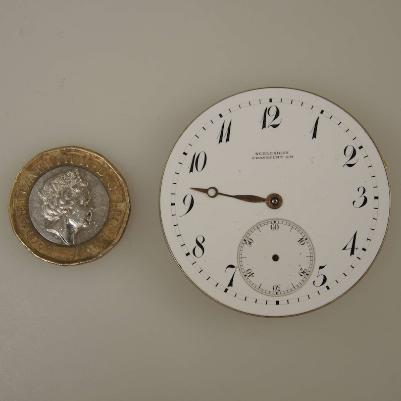 May include: A white pocket watch face with black Roman numeral hour markers and black minute markers. The watch face has a small, white sub-dial with black minute markers. The watch face also has the text "SCHLESICKY FRANKFURT AM" printed on it.