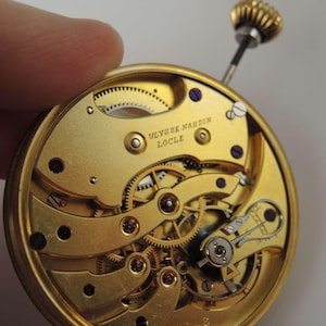 May include: A close-up view of a gold-colored watch movement with intricate gears and a winding stem. The movement is labeled "ULYSSE NARDIN LOCLE".