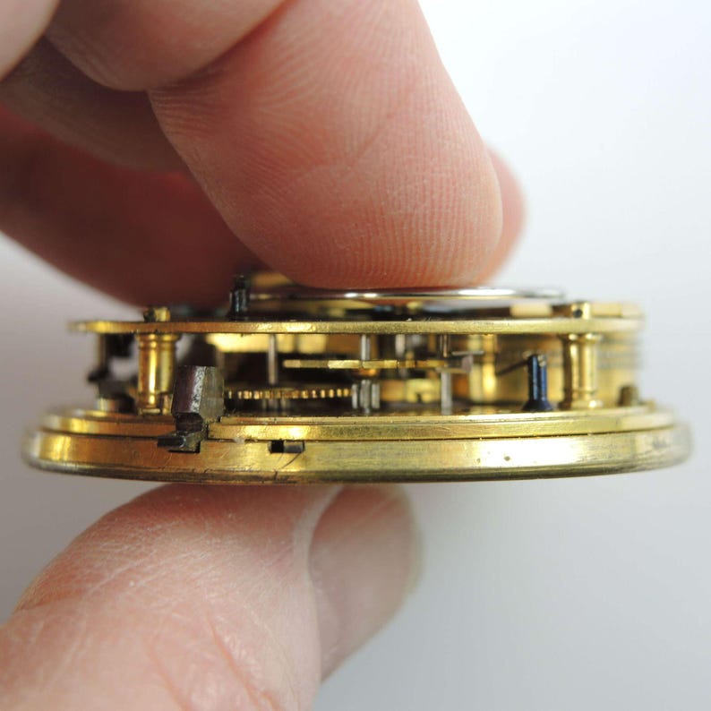 May include: Close-up of a vintage watch movement, showcasing intricate gears and components. The watch mechanism is primarily gold-toned, with visible details of the internal workings. The image highlights the precision engineering of the antique timepiece.