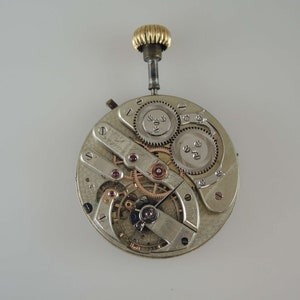 May include: A silver pocket watch movement with a gold winding stem. The watch is disassembled, showing the gears and other internal components.
