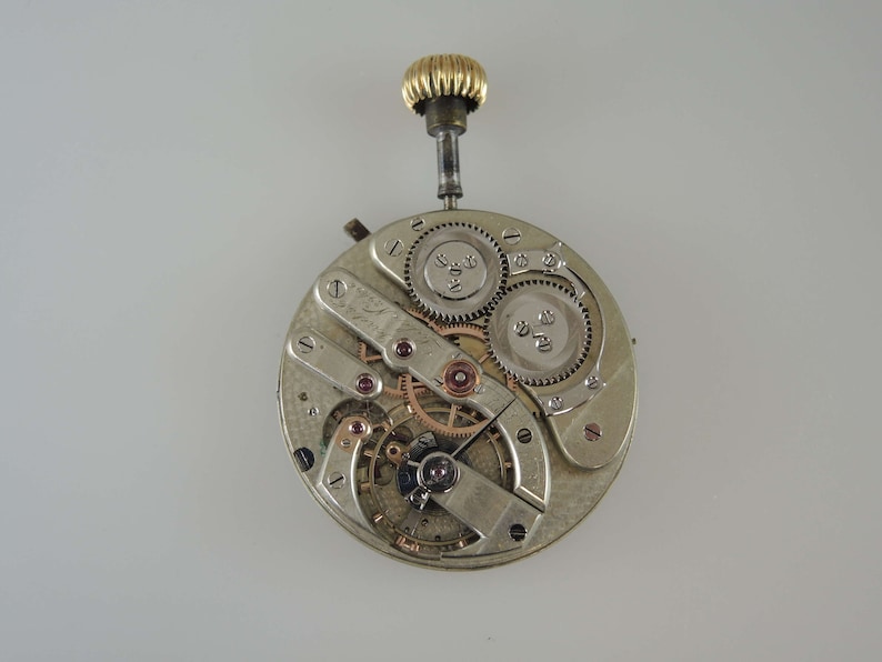 May include: A silver pocket watch movement with a gold winding stem. The watch is disassembled, showing the gears and other internal components.