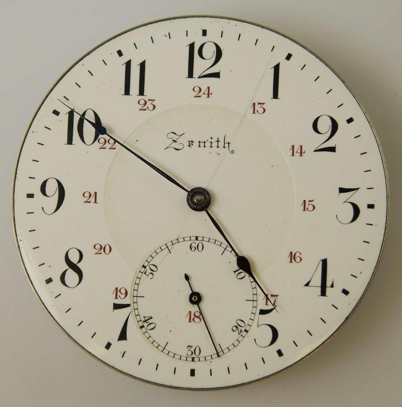 May include: A vintage Zenith pocket watch face with a white background. Black and red numerals mark the hours, with a smaller seconds dial at the bottom. The watch face features the brand name "Zenith" in elegant script.