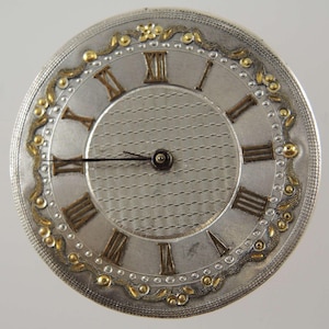 May include: A silver pocket watch face featuring Roman numerals and gold embellishments. The watch face has a textured central area and a detailed outer ring with gold floral patterns. The hands are black, and the design is ornate and vintage.