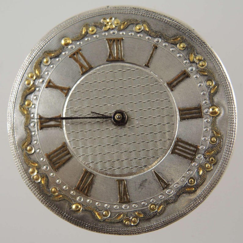 May include: A silver pocket watch face featuring Roman numerals and gold embellishments. The watch face has a textured central area and a detailed outer ring with gold floral patterns. The hands are black, and the design is ornate and vintage.