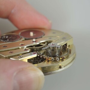 Puede incluir: Primer plano de un movimiento de reloj de tono dorado con engranajes intrincados y un mecanismo de cuerda. El reloj est&aacute; desmontado y se pueden ver las partes internas.