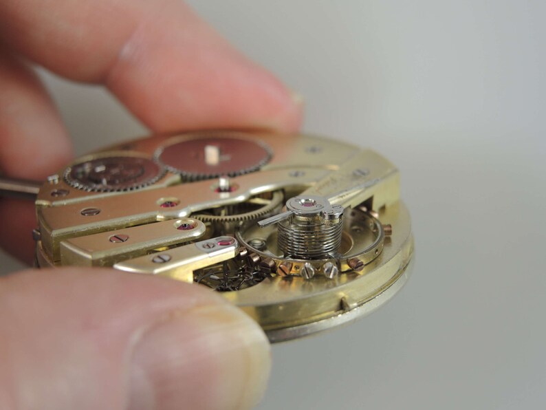 Puede incluir: Primer plano de un movimiento de reloj de tono dorado con engranajes intrincados y un mecanismo de cuerda. El reloj est&aacute; desmontado y se pueden ver las partes internas.