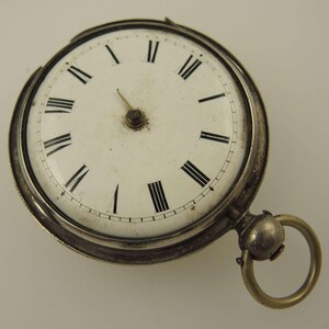 May include: Antique pocket watch with a white face and black Roman numerals. The watch has a silver-colored casing and a gold-colored ring for attaching a chain. The watch face is slightly aged, adding to its vintage appeal.