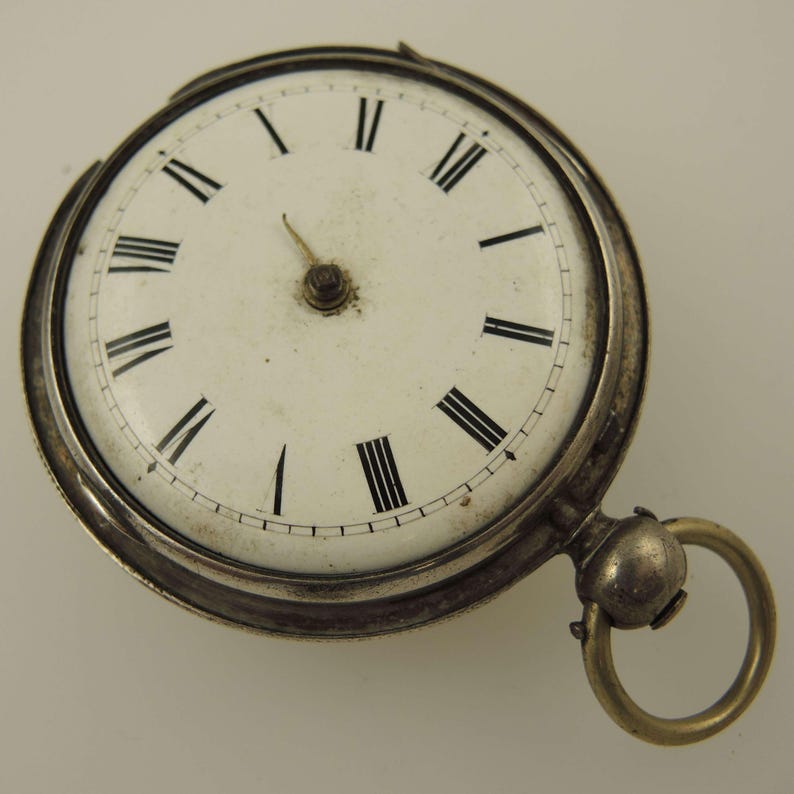 May include: Antique pocket watch with a white face and black Roman numerals. The watch has a silver-colored casing and a gold-colored ring for attaching a chain. The watch face is slightly aged, adding to its vintage appeal.